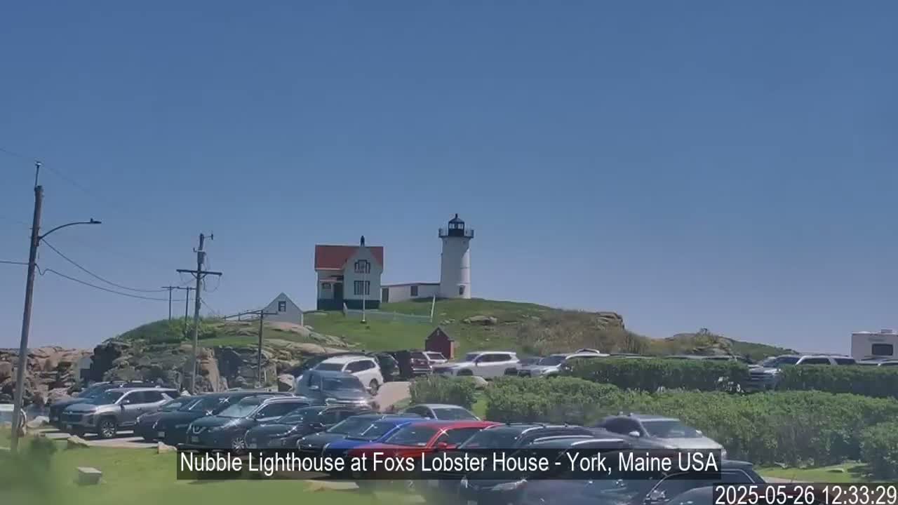 A lighthouse sits atop a grassy hill overlooking a large parking lot full of cars under a clear blue sky.