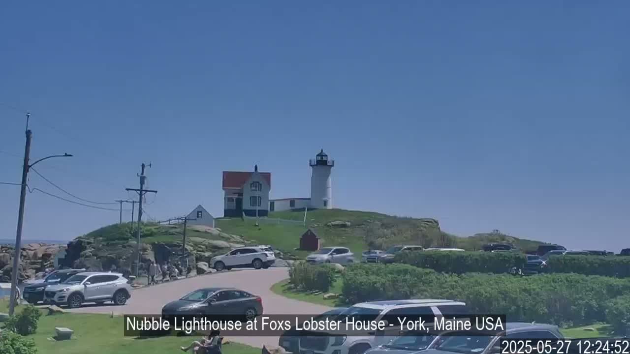A lighthouse and keeper's house sit atop a rocky hill overlooking a parking lot filled with cars on a sunny day.