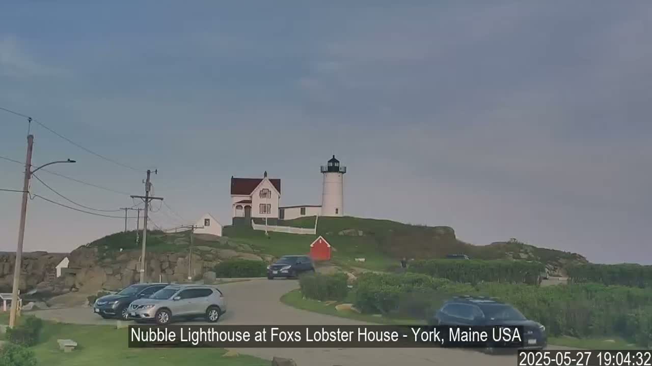 A white lighthouse and a smaller white house sit atop a rocky hill overlooking a parking lot with several cars under a mostly clear sky.