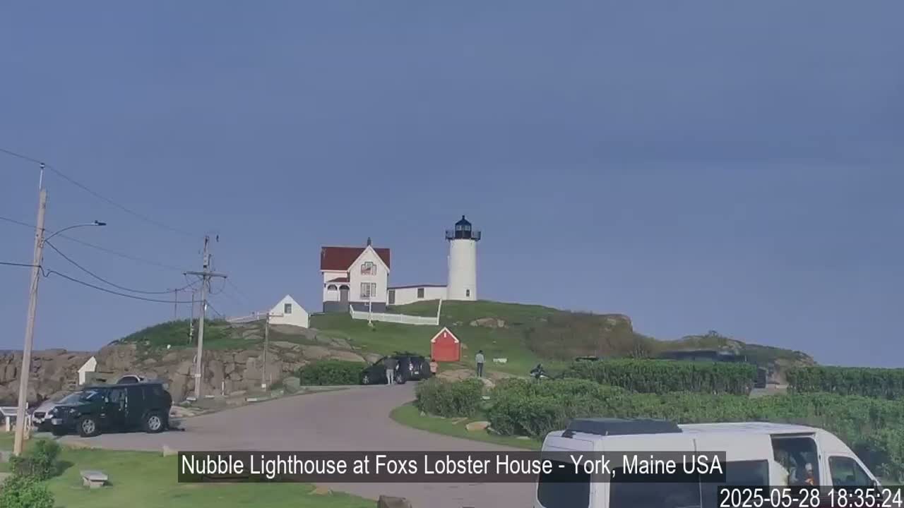 A white lighthouse and keeper's house sit atop a rocky hill overlooking a parking lot with several vehicles under a clear blue sky.