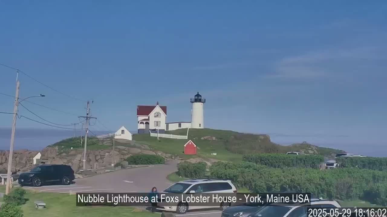 A white lighthouse sits atop a rocky hill overlooking a calm ocean under a clear blue sky, with several cars parked in a nearby lot.