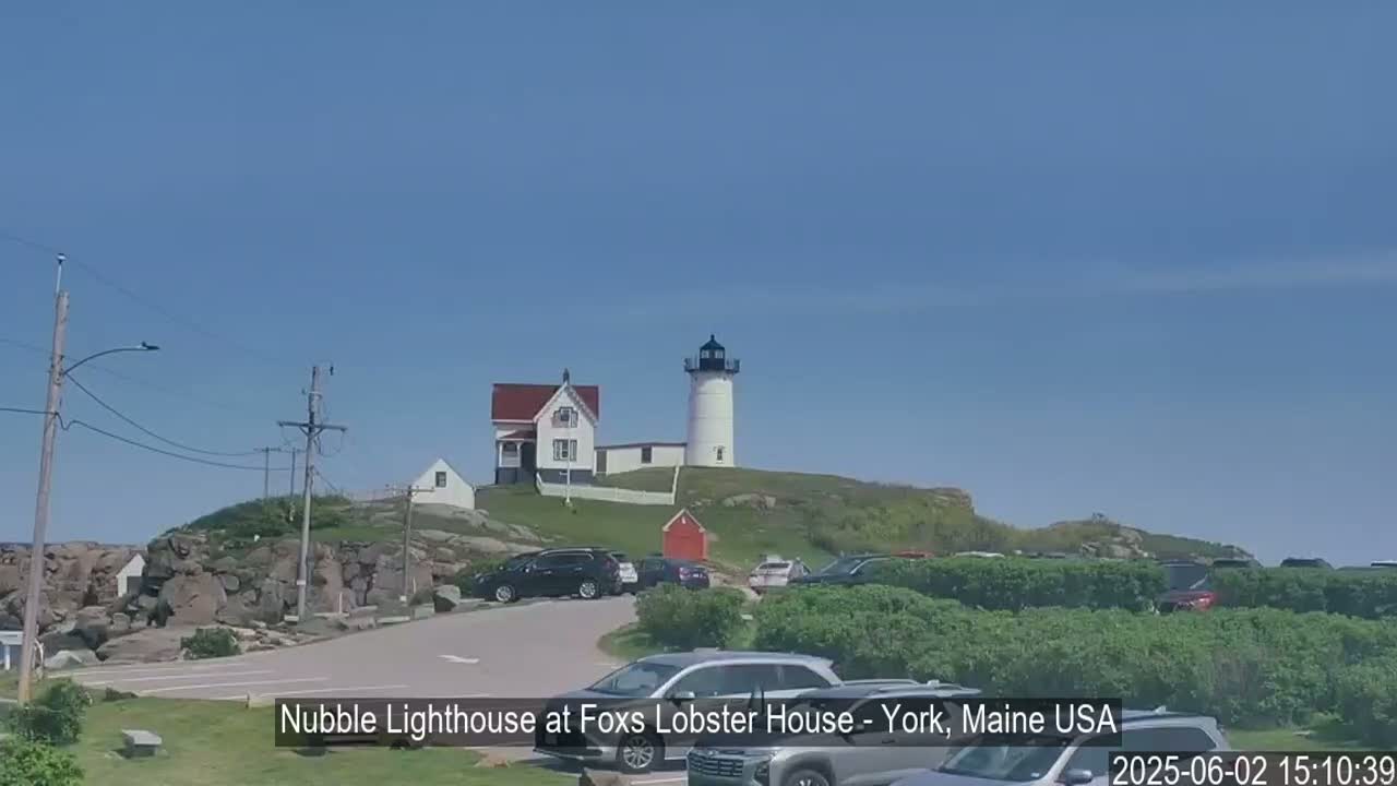 A lighthouse sits atop a rocky hill overlooking a parking lot filled with cars on a clear, sunny day.