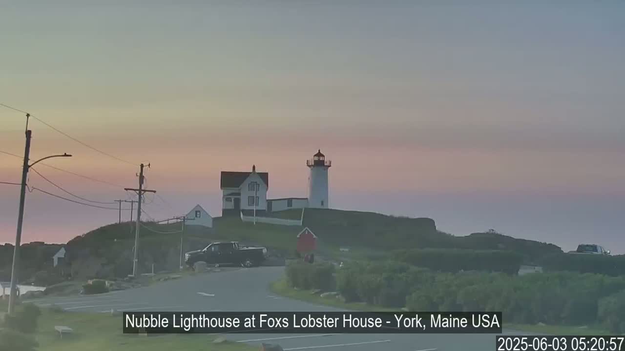 A lighthouse and accompanying buildings sit atop a small hill at dawn under a pale pink and gray sky.
