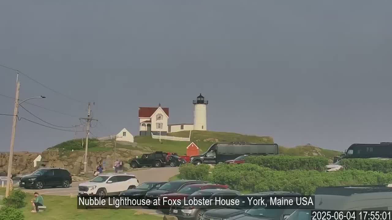 A lighthouse sits atop a rocky hill overlooking a parking lot filled with cars and buses under an overcast sky.