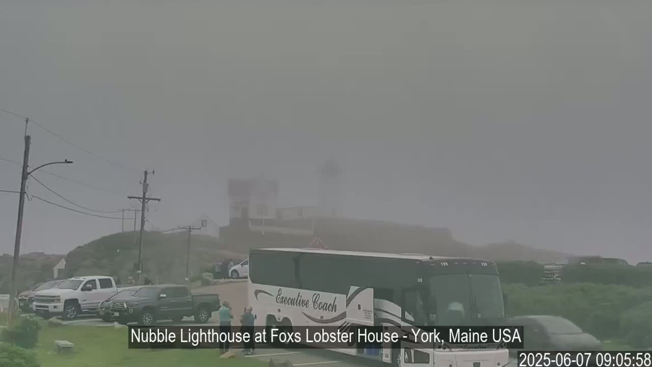 A foggy day reveals a lighthouse in the distance, with several cars and a tour bus parked in a lot in the foreground.