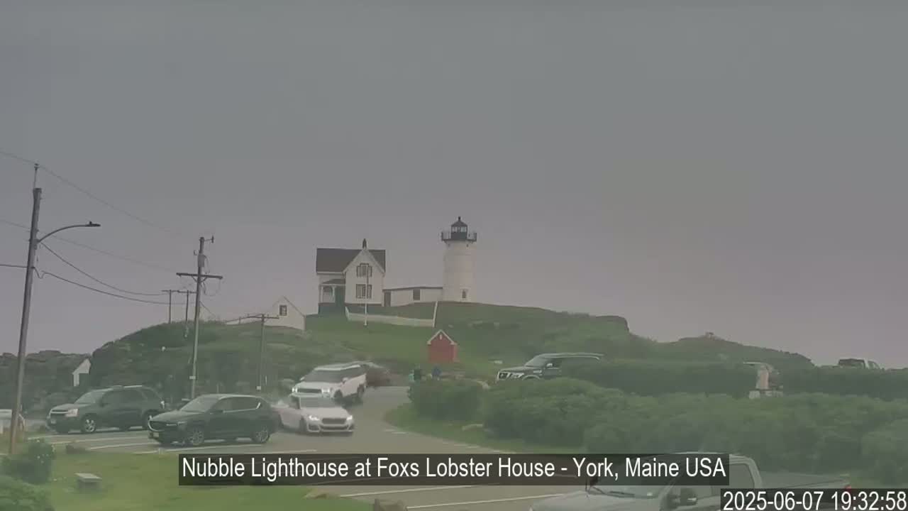 A lighthouse sits atop a hill with several cars parked nearby on an overcast day.