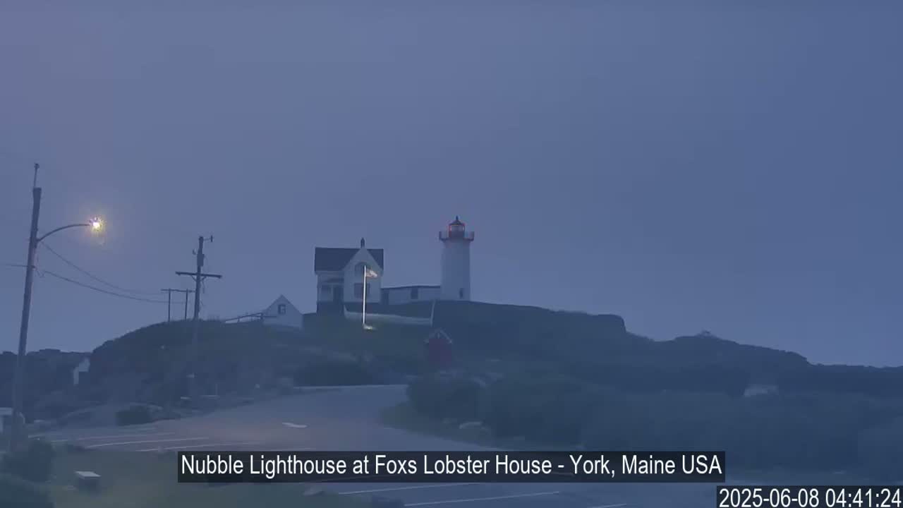 A lighthouse and a smaller building sit atop a dark hill under a hazy, light-blue sky.