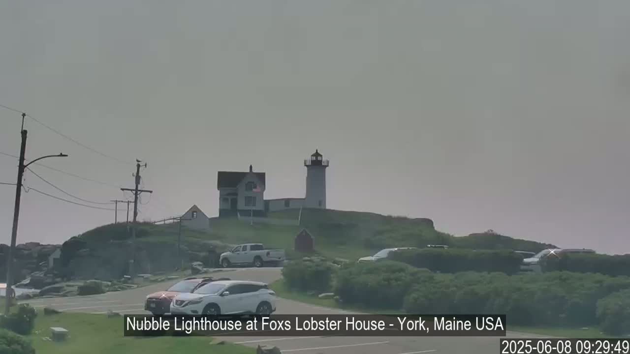 A lighthouse and accompanying buildings sit atop a hill overlooking a parking lot with several cars, under a hazy sky.