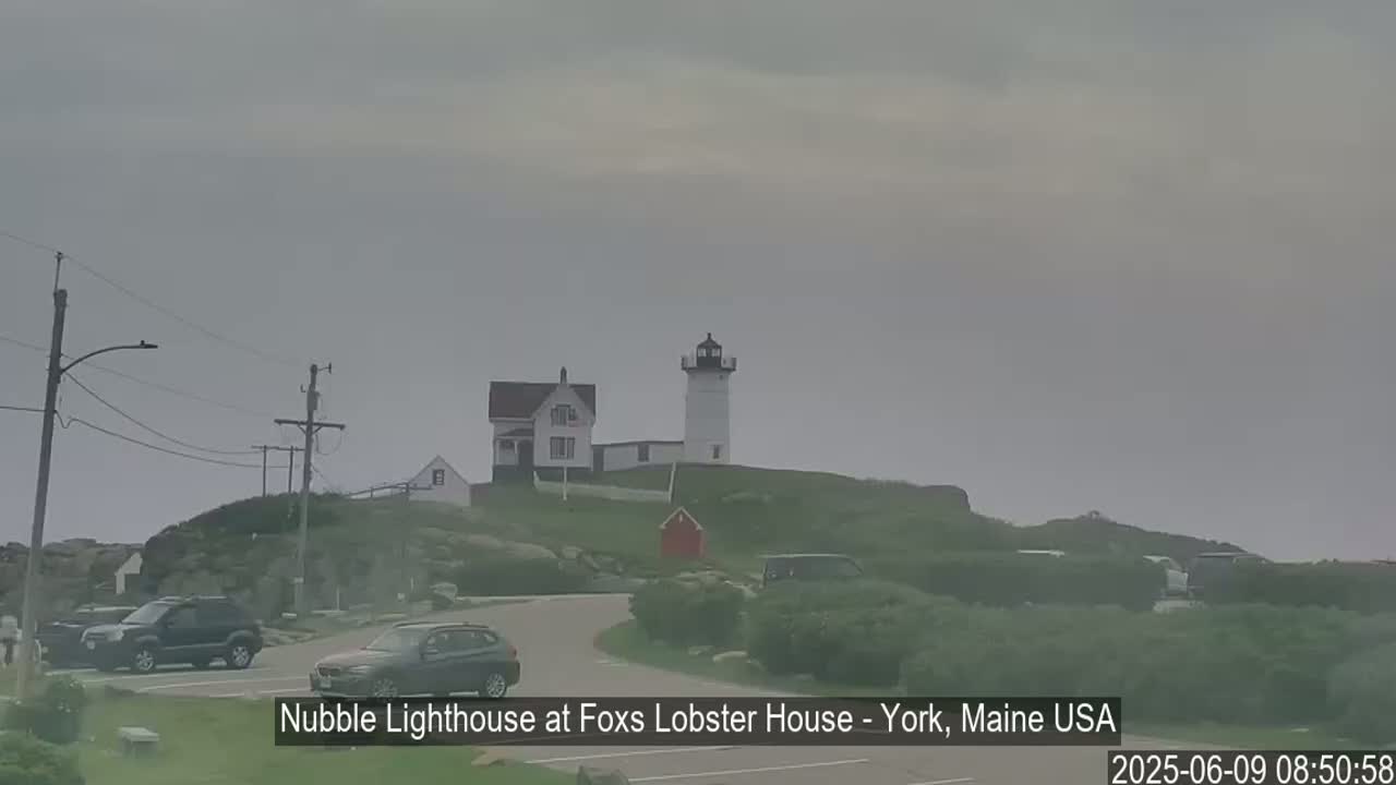 A lighthouse and a smaller building sit atop a rocky hill overlooking a parking lot with several cars under an overcast sky.