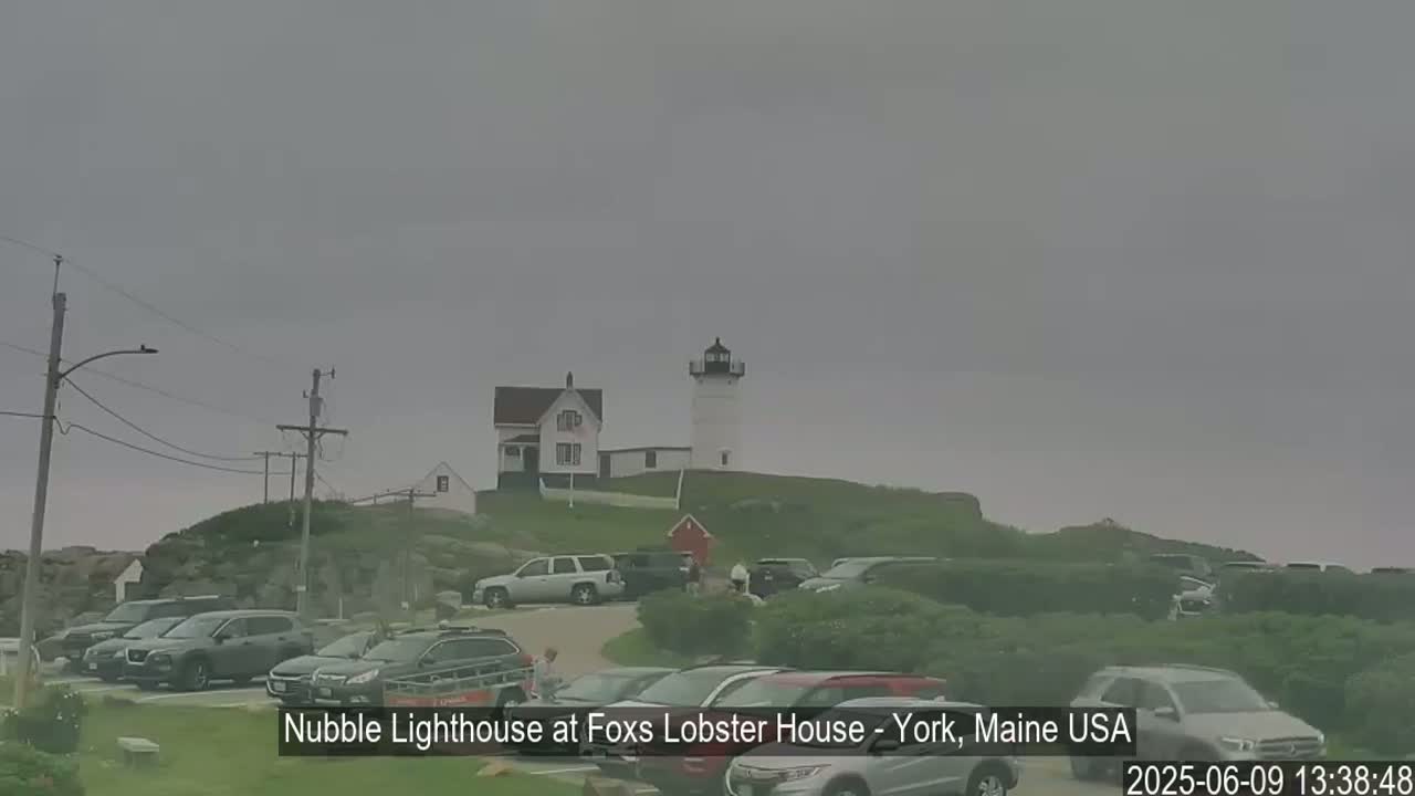A lighthouse and several cars are visible on a cloudy day, with some people near the cars.