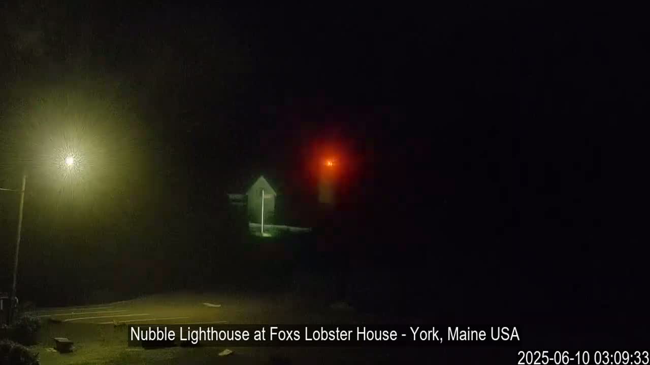 A lighthouse with a red light shines faintly through a foggy, nighttime sky, illuminated by a nearby streetlight.