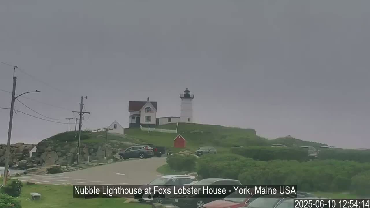 A lighthouse and keeper's house sit atop a rocky hill on an overcast day, with several cars parked nearby.