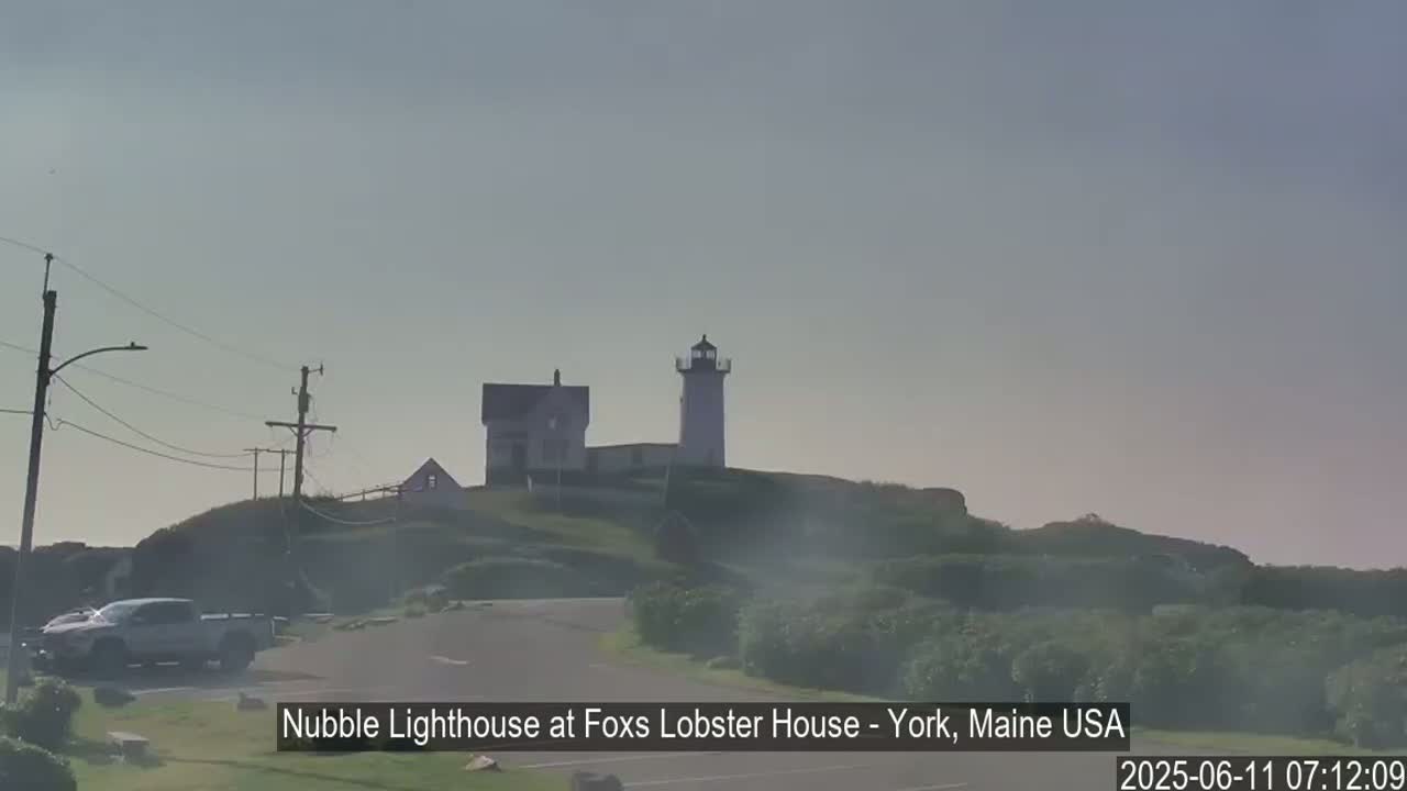 A lighthouse and a building sit atop a hill covered in low-lying vegetation under a hazy sky.
