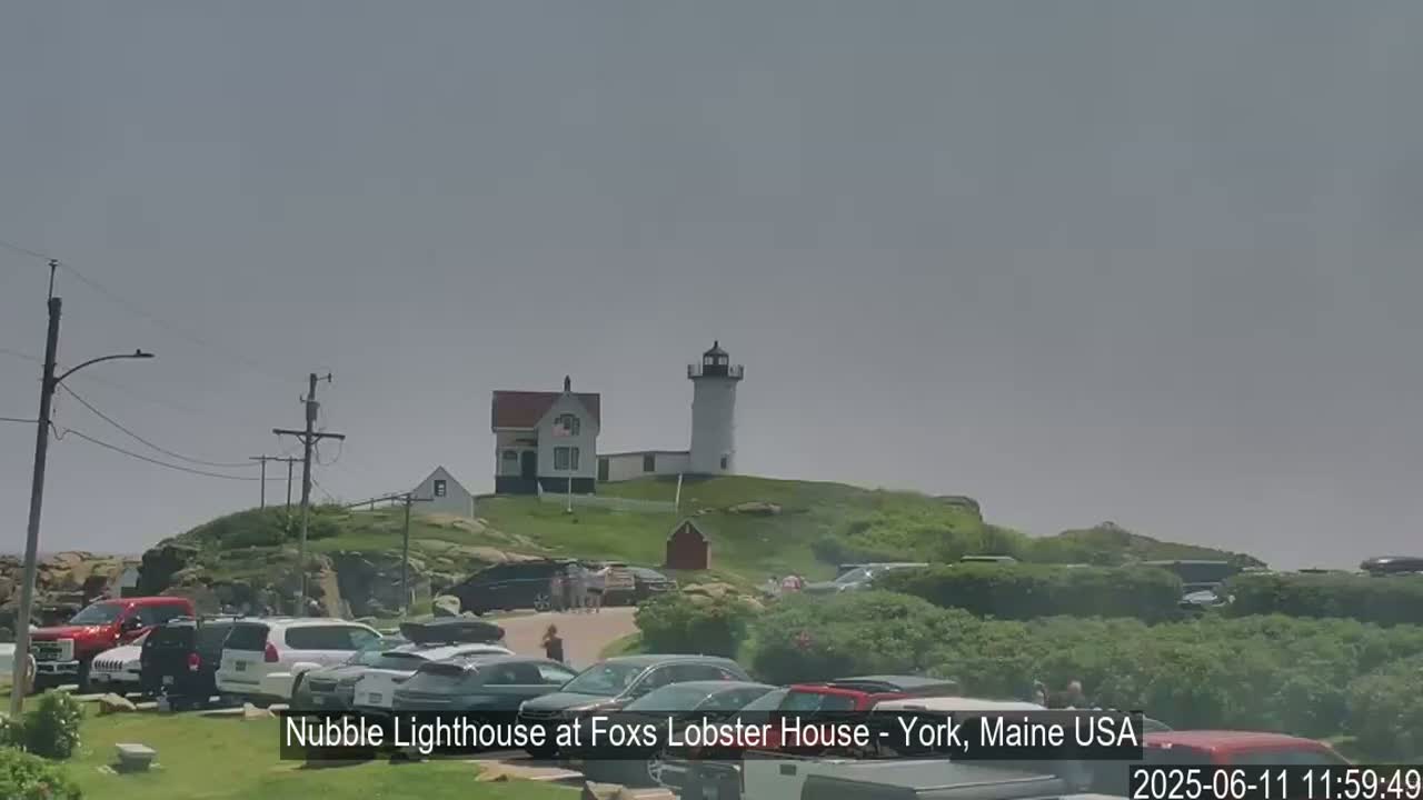 A lighthouse and a smaller building sit atop a grassy hill overlooking a parking lot filled with cars on a hazy day.