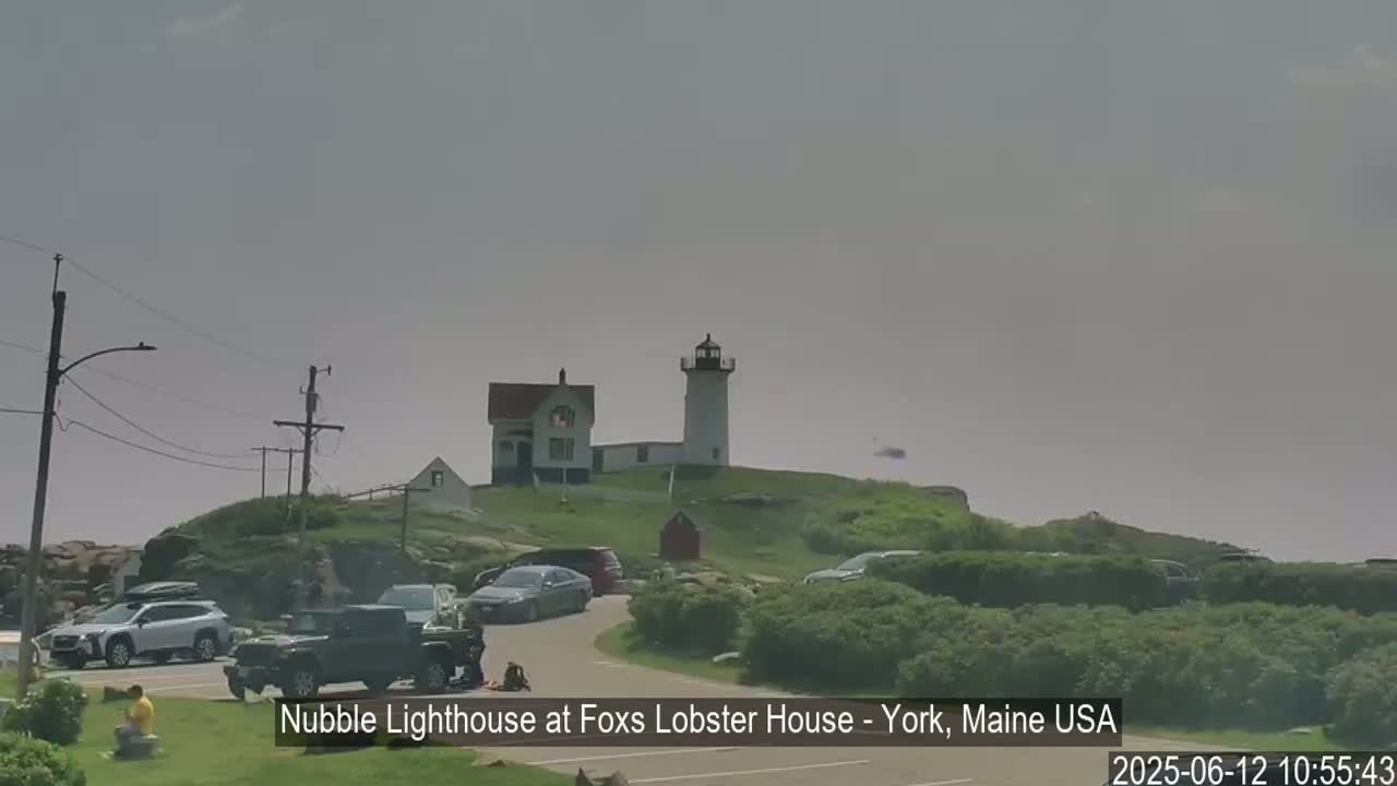 A lighthouse sits atop a grassy hill overlooking a parking lot filled with cars on an overcast day.