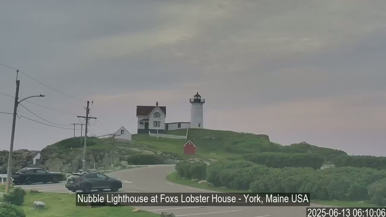 A lighthouse and keeper's house sit atop a grassy hill overlooking a road with parked cars under an overcast sky.