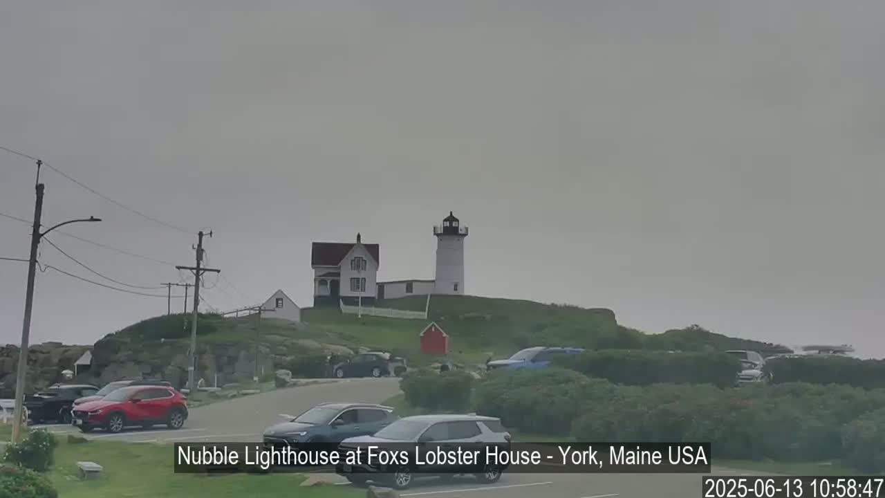 A lighthouse and a nearby house sit atop a grassy hill overlooking a parking lot filled with cars on an overcast day.