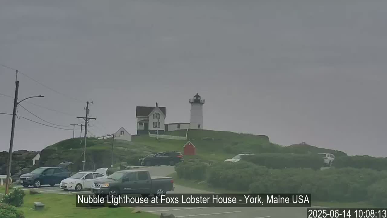 A lighthouse and accompanying buildings sit atop a grassy hill on an overcast day, with several cars parked in a lot below.
