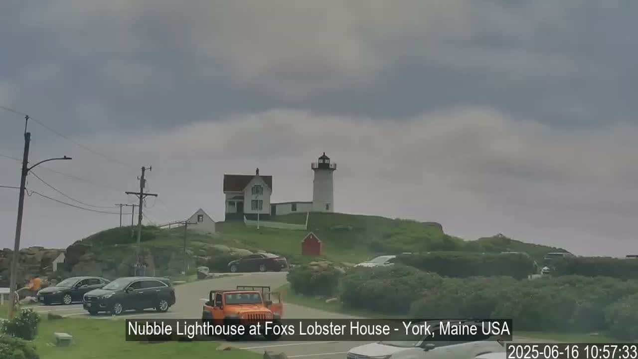 A lighthouse and keeper's house sit atop a grassy hill overlooking a parking lot with several cars, under an overcast sky.
