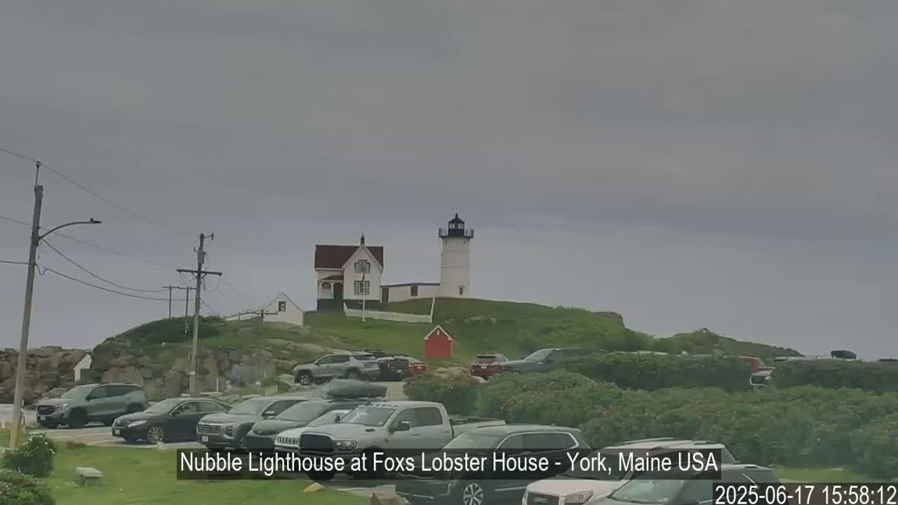A white lighthouse sits atop a grassy hill overlooking a parking lot full of cars on an overcast day.