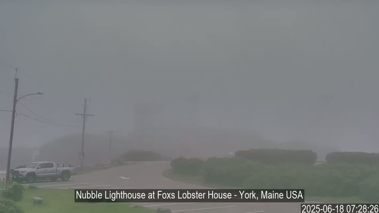 A foggy scene shows a parking lot with a few vehicles, utility poles, and low-lying vegetation near a road leading towards a barely visible structure in the distance.