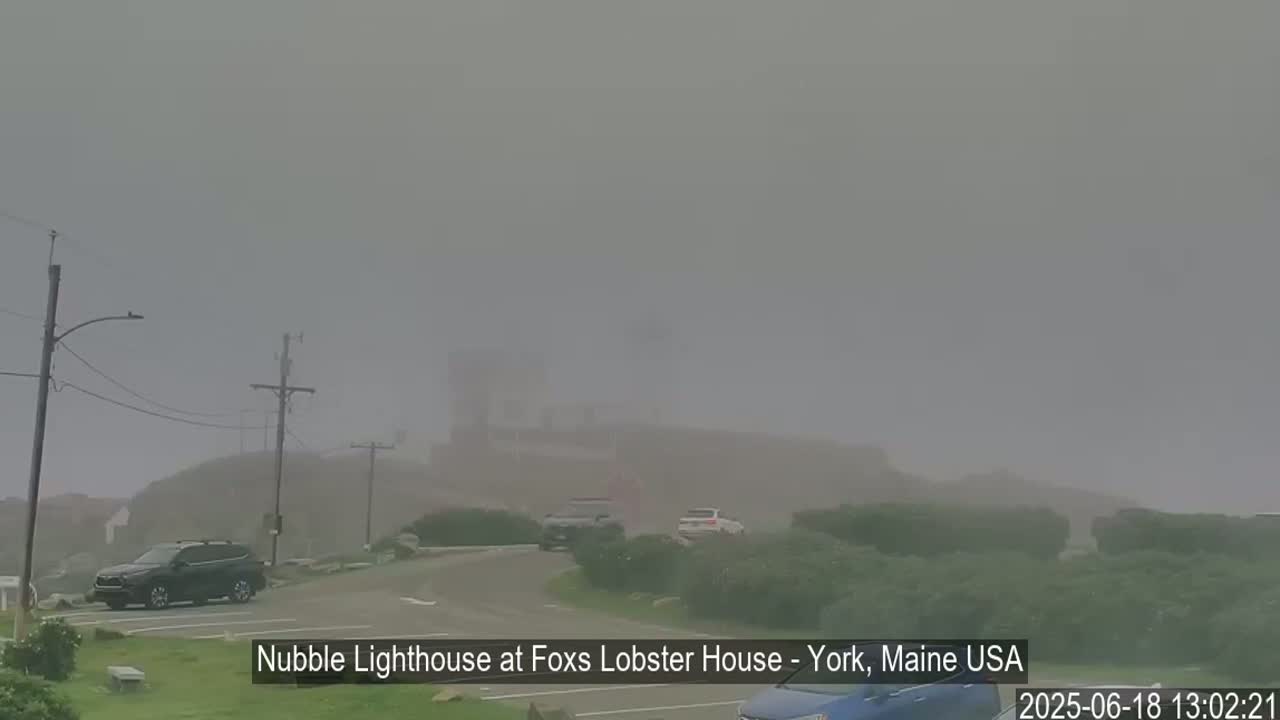 A lighthouse is barely visible through a thick fog, with a few cars parked near a road in the foreground.