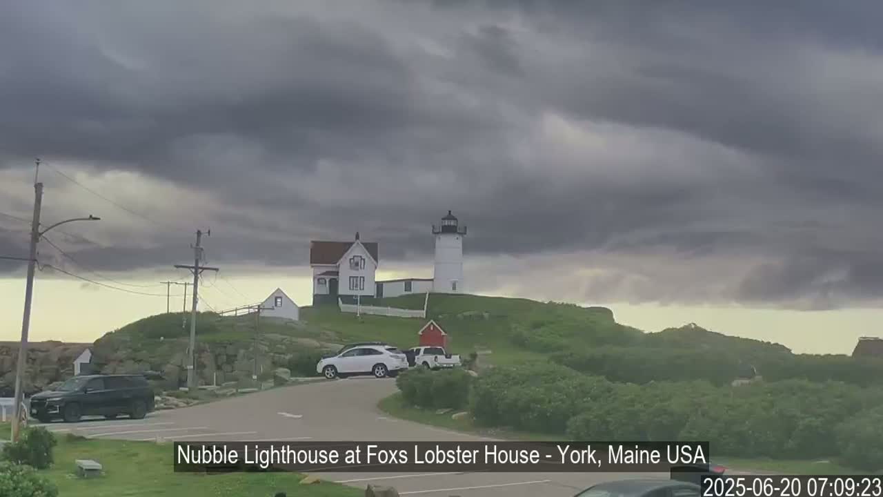 A lighthouse sits atop a grassy hill under a dark, cloudy sky, with several cars parked nearby in a parking lot.