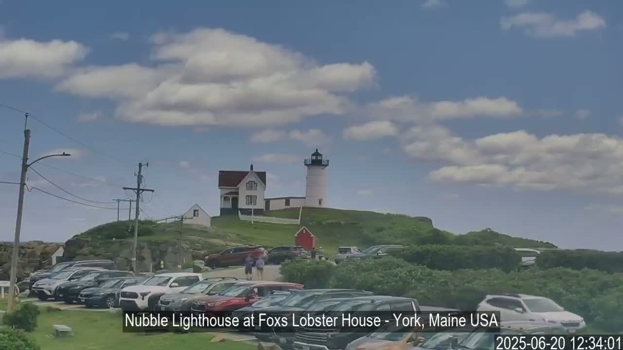 A lighthouse and keeper's house sit atop a grassy hill overlooking a parking lot filled with cars under a partly cloudy sky.