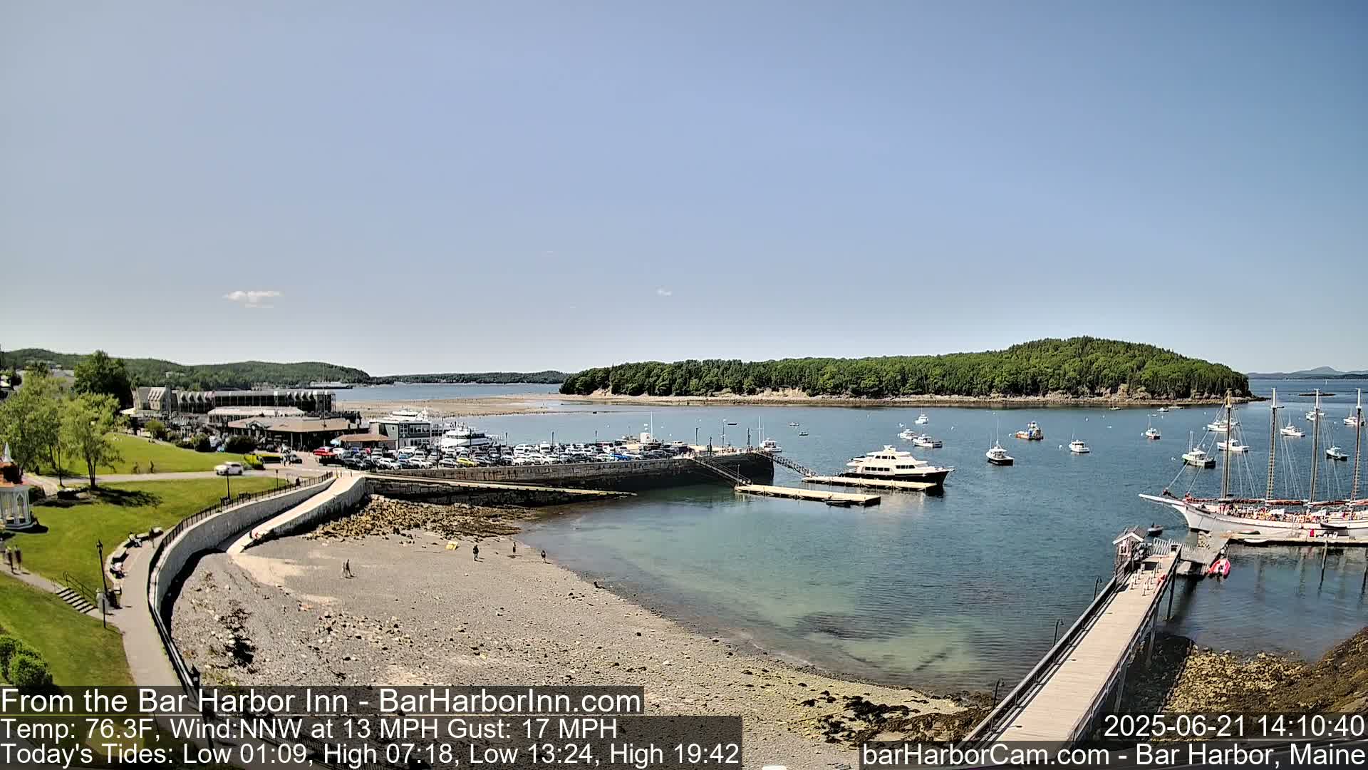 A sunny harbor scene shows a rocky beach, a parking lot, several boats and a pier, with a wooded island in the distance.