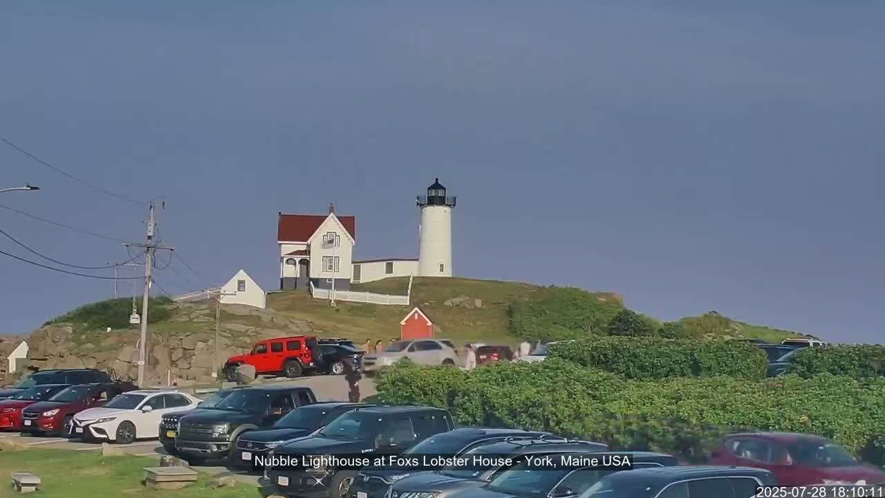 A lighthouse and keeper's house sit atop a rocky hill overlooking a parking lot filled with cars on a clear day.