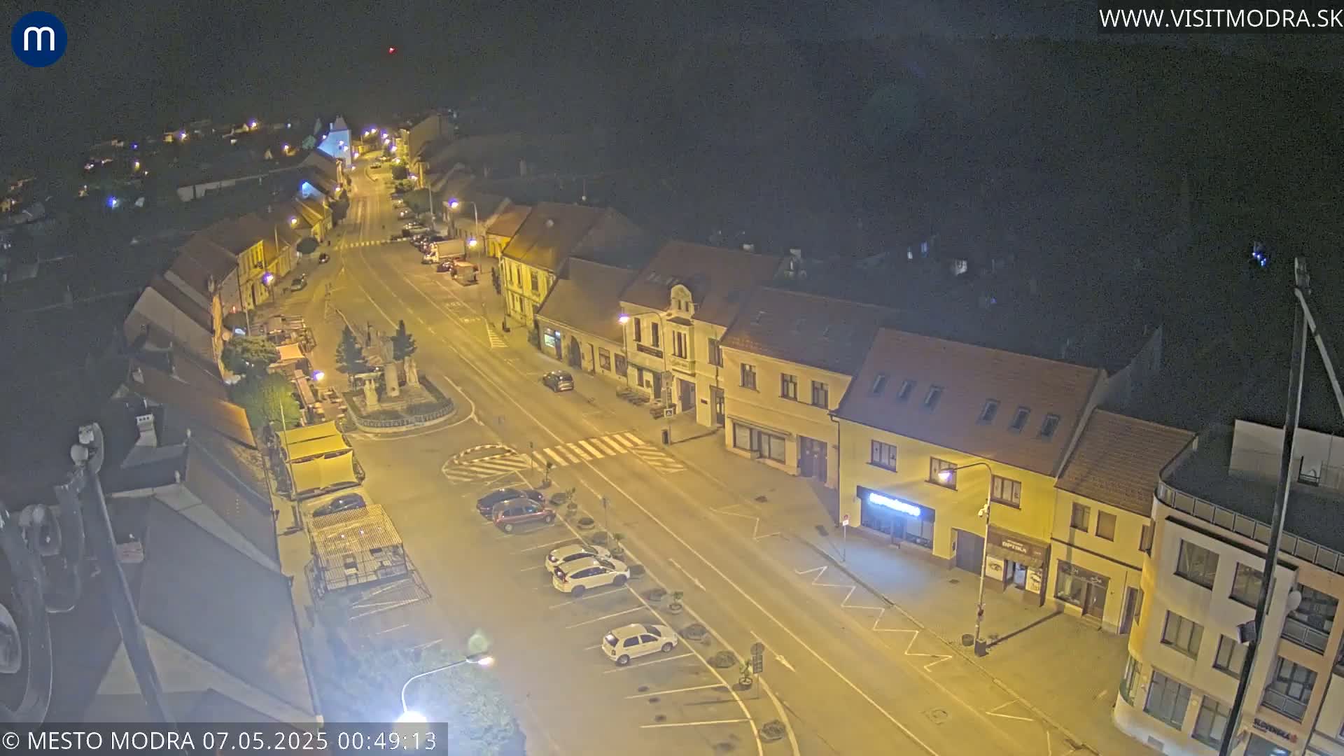 A nighttime, high-angle view of a town's street scene showing parked cars and buildings illuminated by streetlights.
