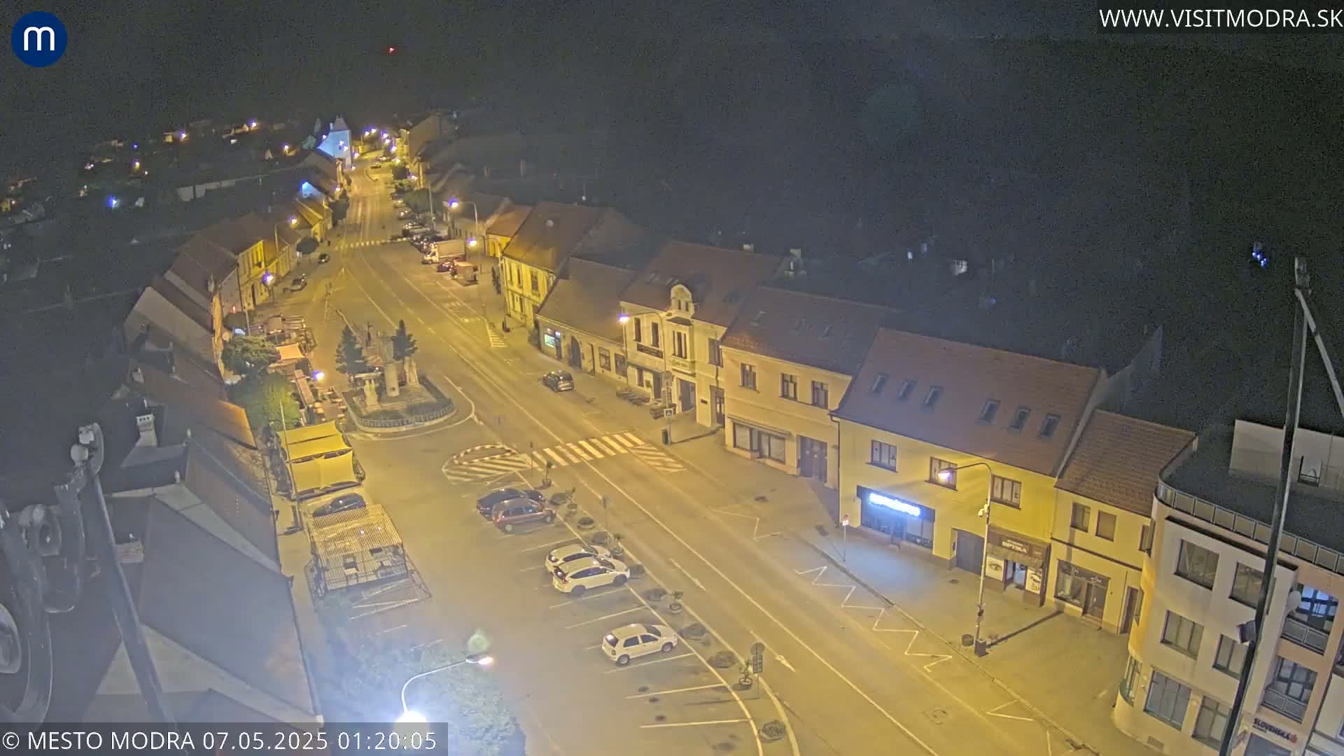 An aerial nighttime view shows a town square with parked cars and buildings lining a street under a clear, dark sky.