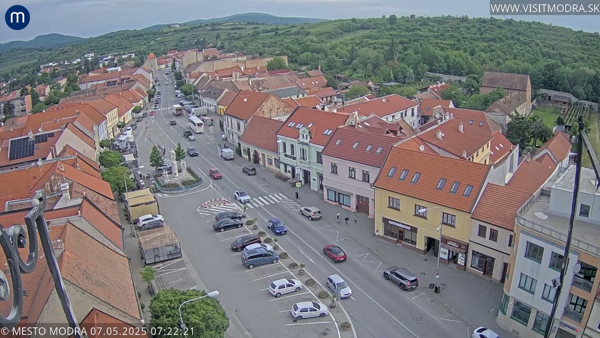 An aerial view shows a town's main street with parked cars and several buildings under an overcast sky.