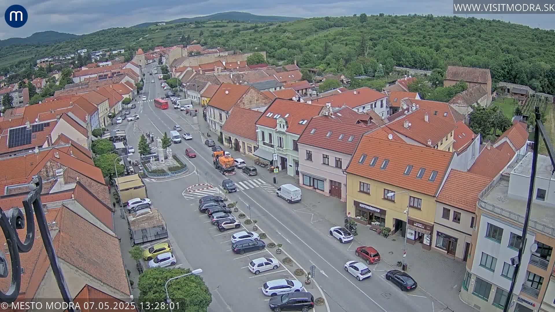 An aerial view of a town's main street, under a partly cloudy sky, shows buildings with red tile roofs, parked cars, and a few vehicles in motion.