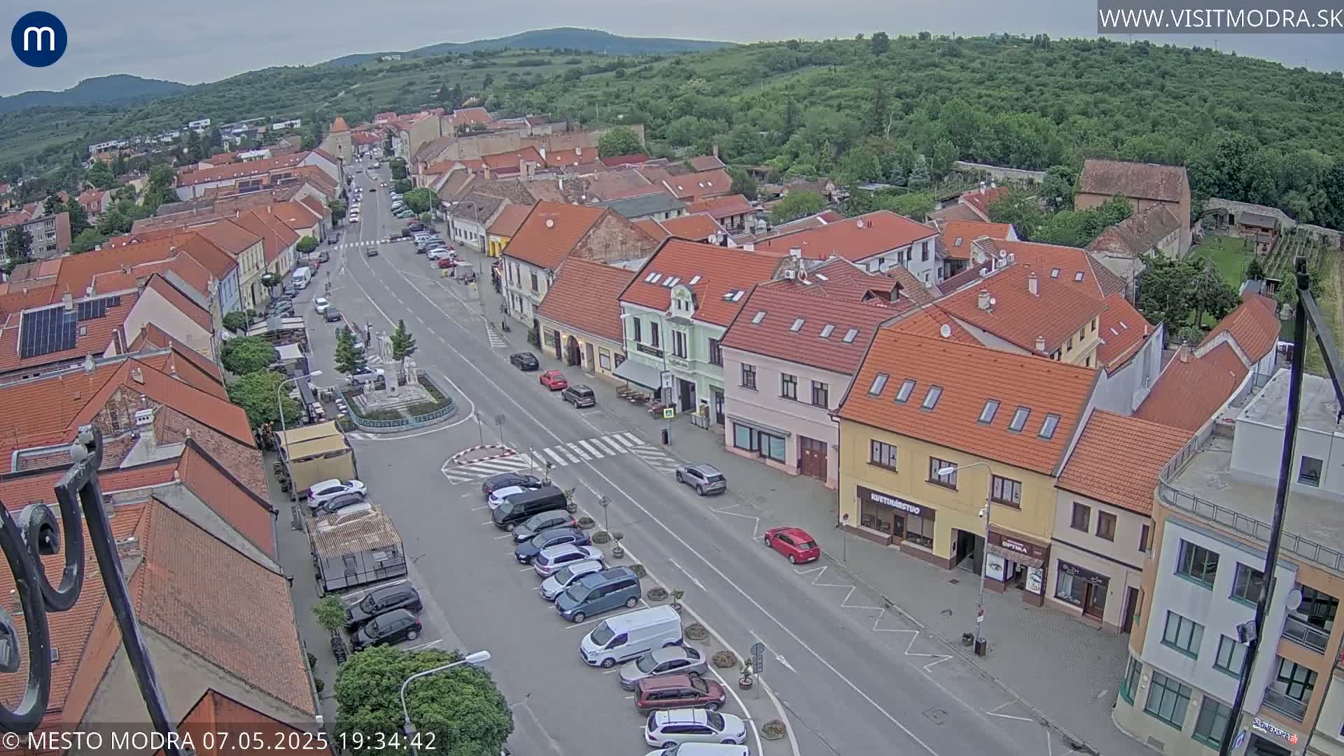 An aerial view shows a town square with parked cars lining a street, surrounded by low-rise buildings under an overcast sky.