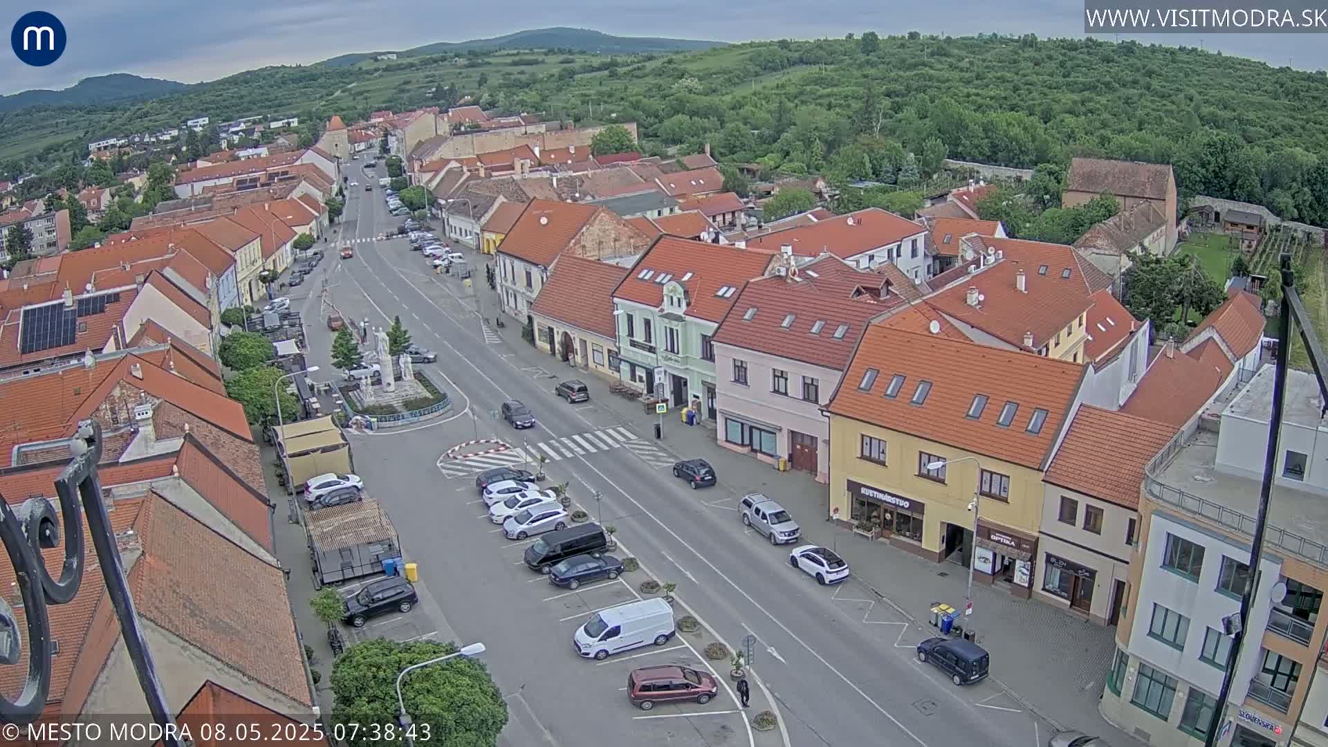 An aerial view of a town square and surrounding buildings on an overcast day, showing parked cars, a pedestrian crossing, and a fountain in the center.