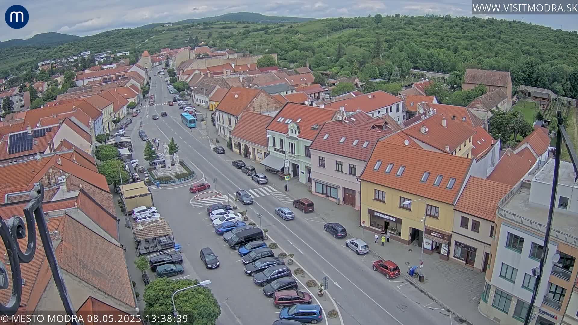An aerial view shows a town square with parked cars lining a street, and red-tiled buildings under a partly cloudy sky.