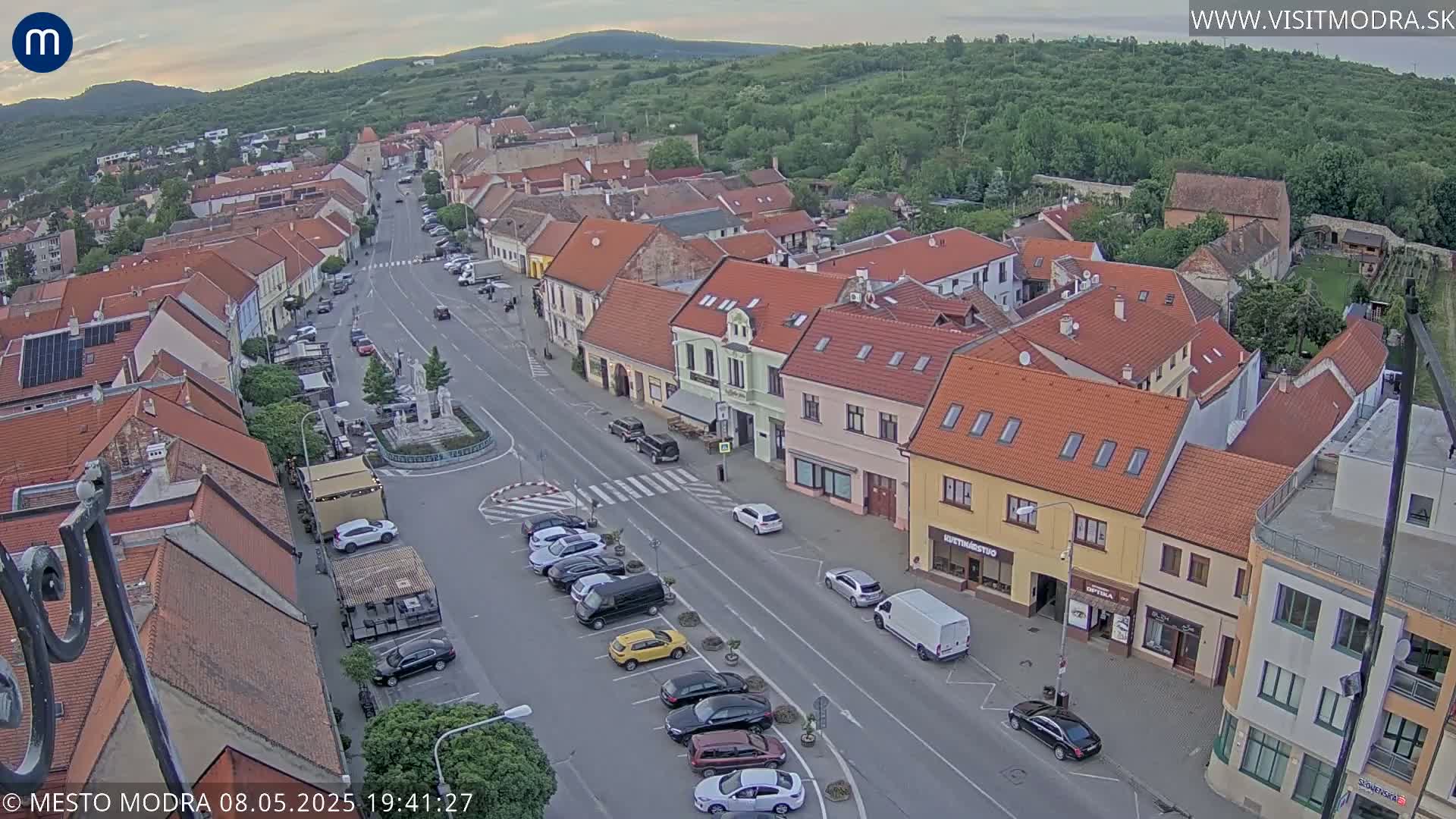 An aerial view of a town square with cars parked along the street and buildings of varying heights and colors under a partly cloudy sky.