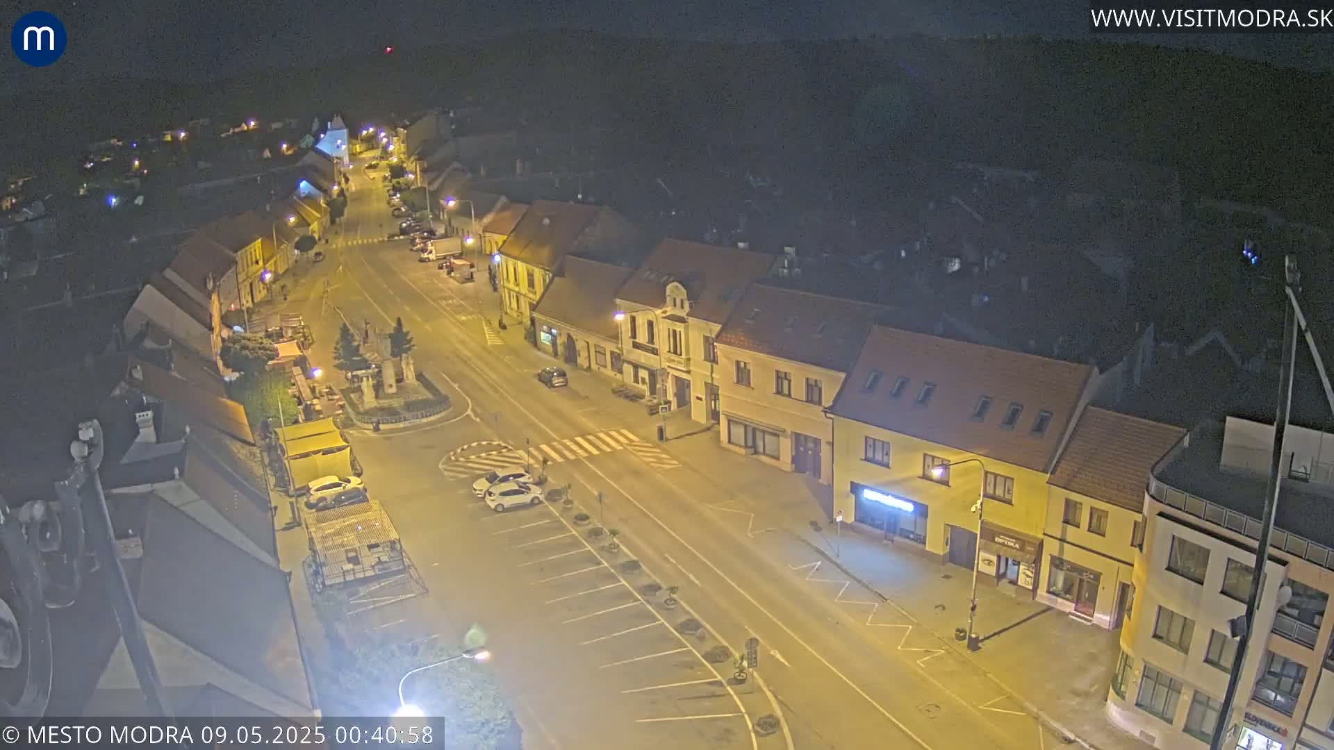 A nighttime, high-angle view shows a town square with several parked cars and buildings illuminated by streetlights.