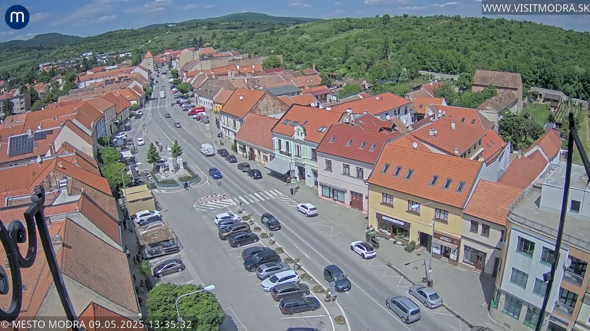 An aerial view of a town square on a sunny day, showing a main street lined with parked cars and buildings with red-tiled roofs, and a fountain in the center.