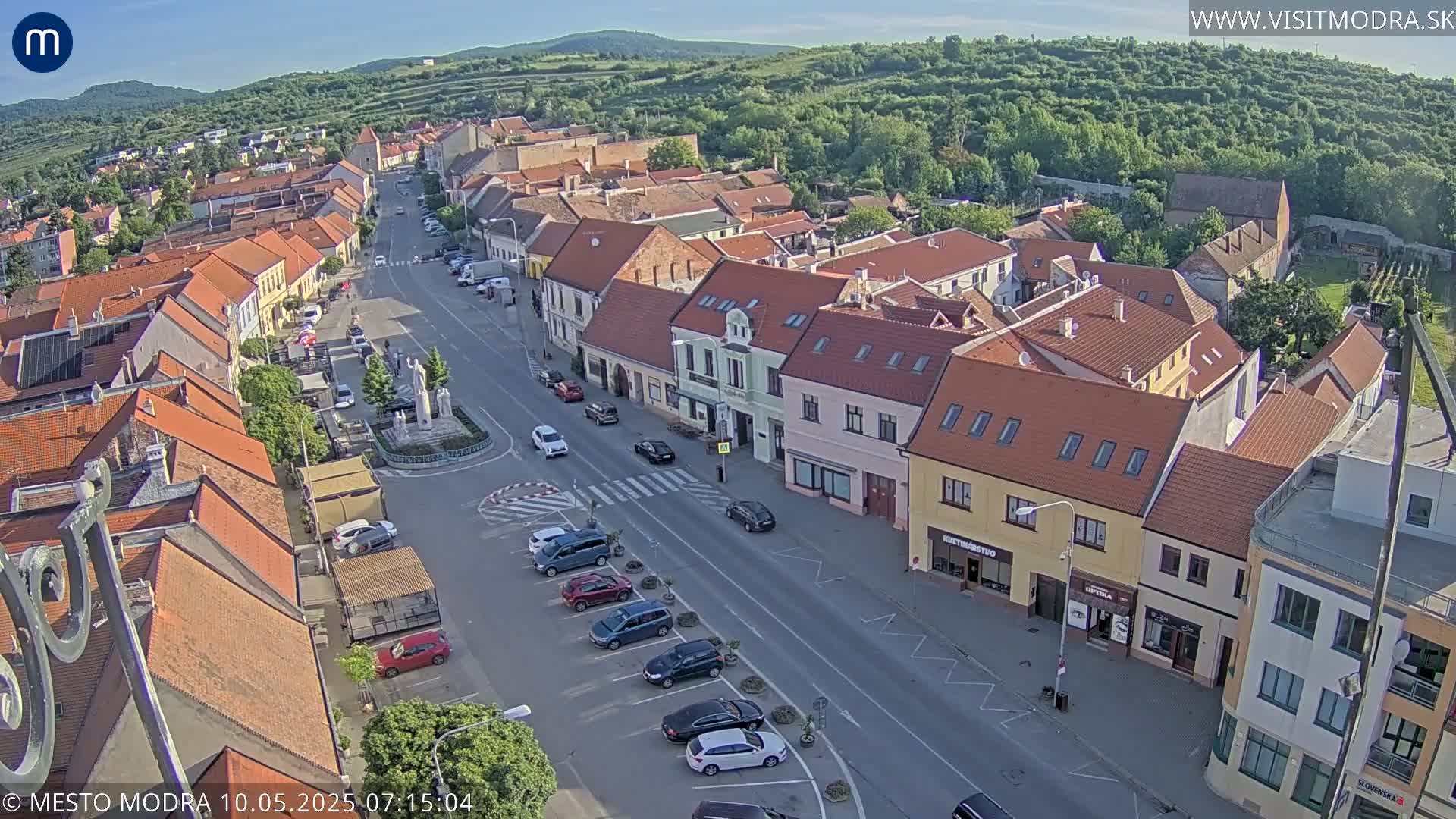 An aerial view shows a town square with parked cars and buildings under a mostly clear sky.