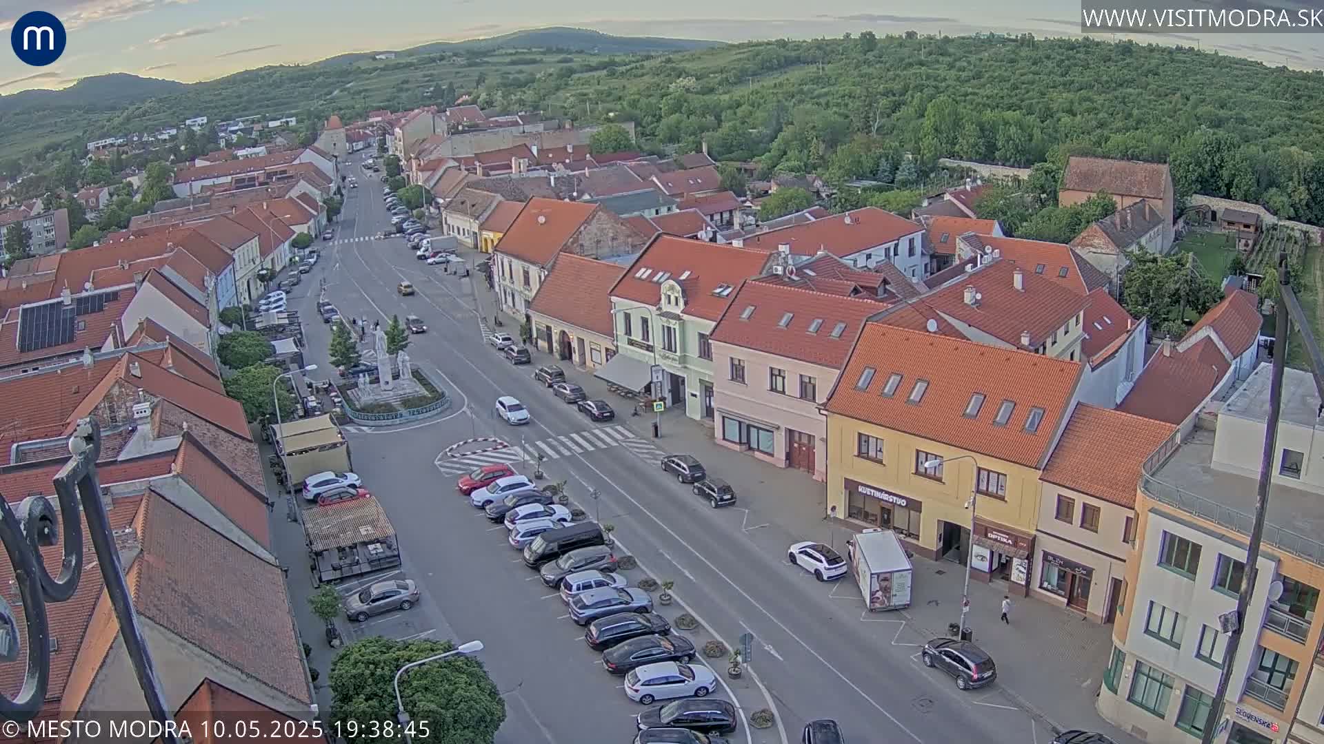 An aerial view of a town square with parked cars lining the street under a partly cloudy evening sky.
