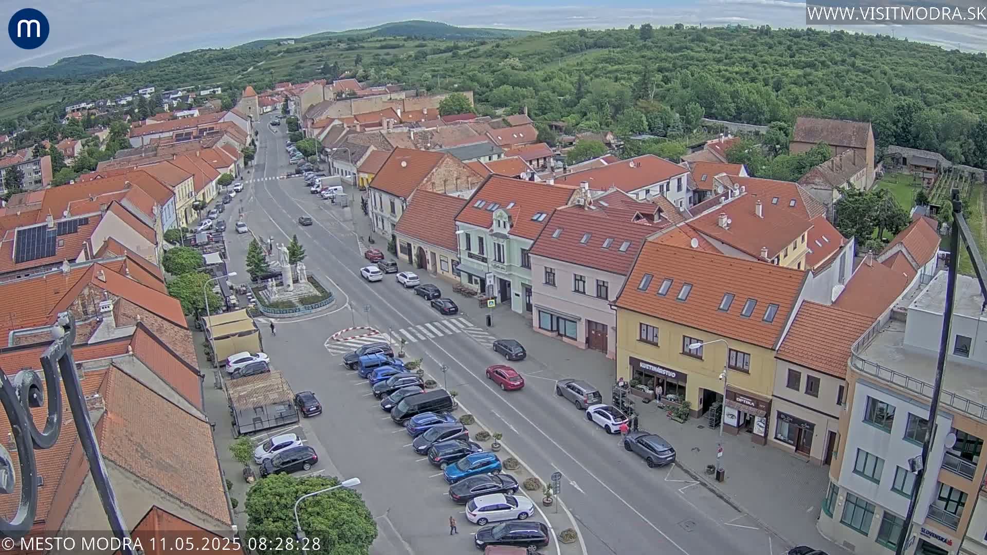 A partly cloudy day reveals a high-angle view of a town square with parked cars lining a main road, surrounded by buildings with red tile roofs and green hills in the background.