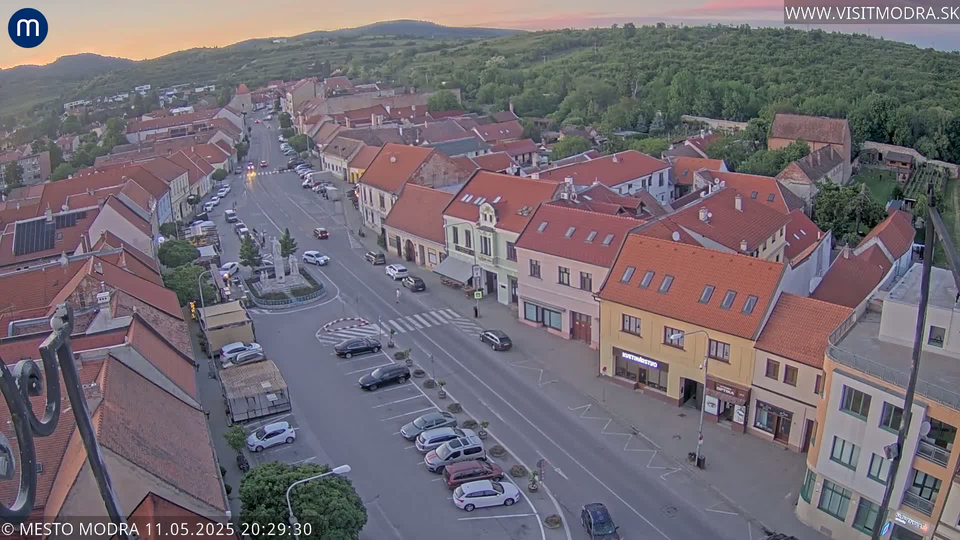 An aerial view shows a town square with parked cars and buildings at sunset under a clear sky.