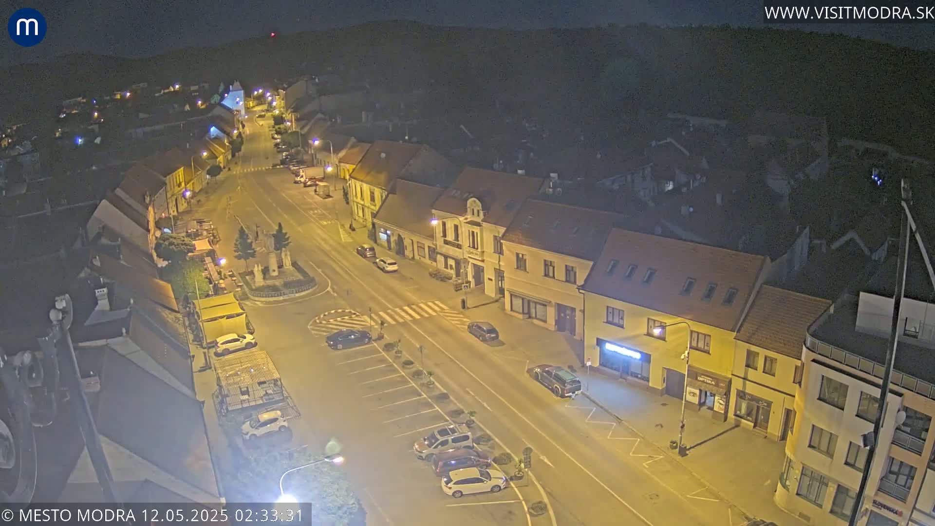 An aerial nighttime view of a town square and surrounding buildings, illuminated by streetlights under a clear, dark sky.