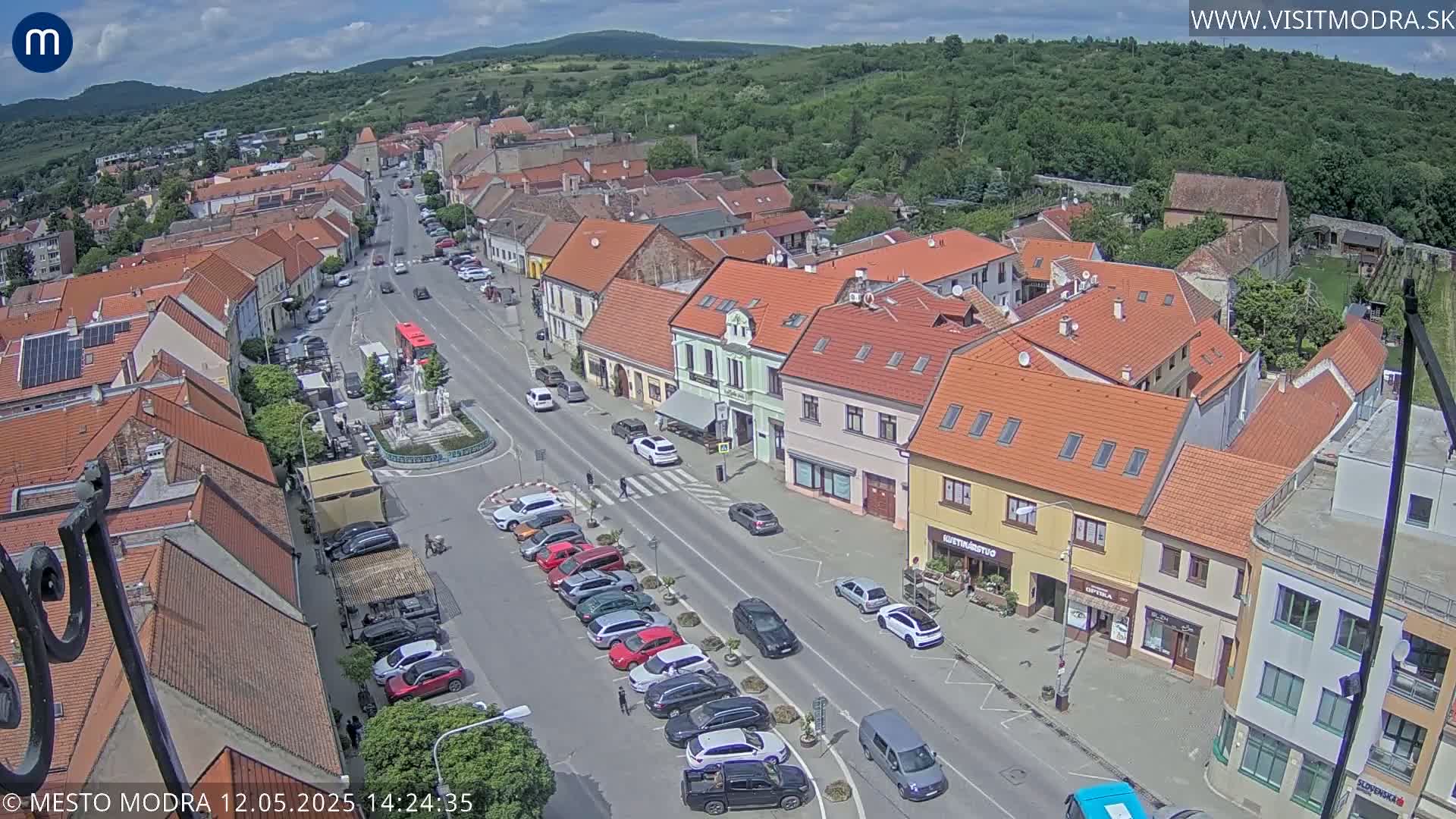 A high-angle, sunny view of a town square with parked cars lining a street in front of buildings with red tile roofs, under a partly cloudy sky.