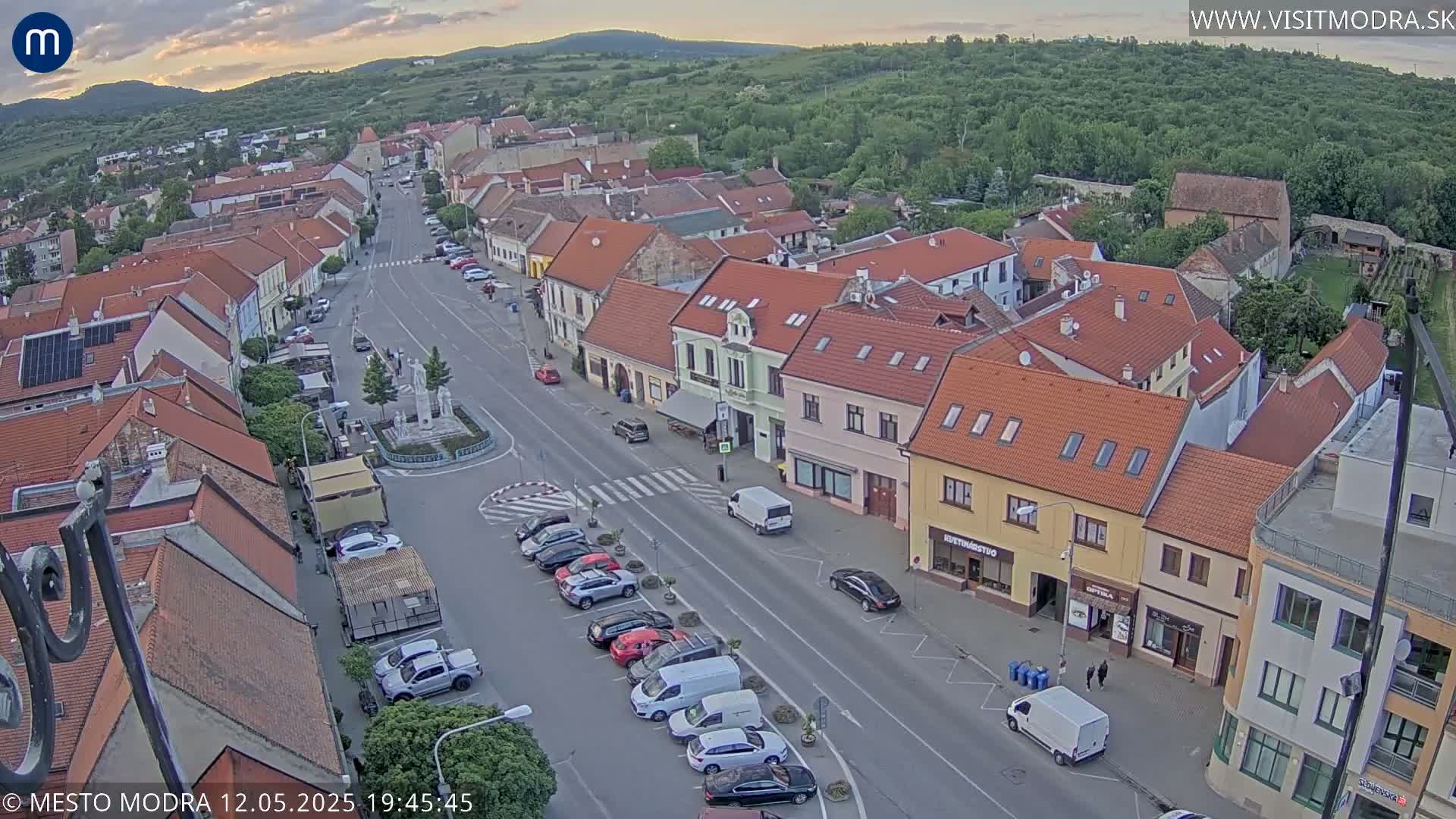 An aerial view of a town square at dusk, showing parked cars lining a main street flanked by low buildings, with clear skies and gentle hills in the background.
