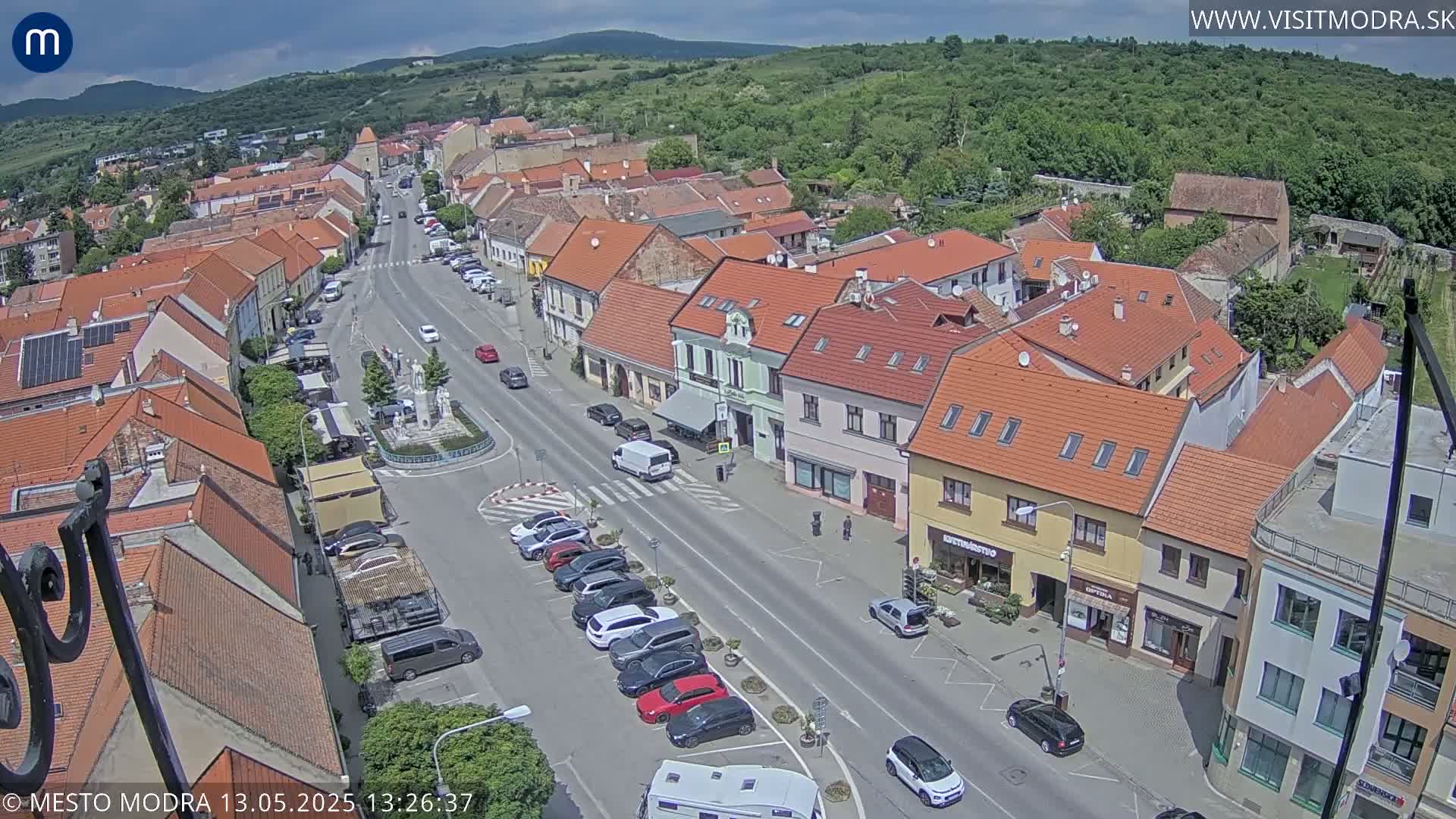 A high-angle view of a town's main street on a partly sunny day, showing parked cars and buildings with red tile roofs.