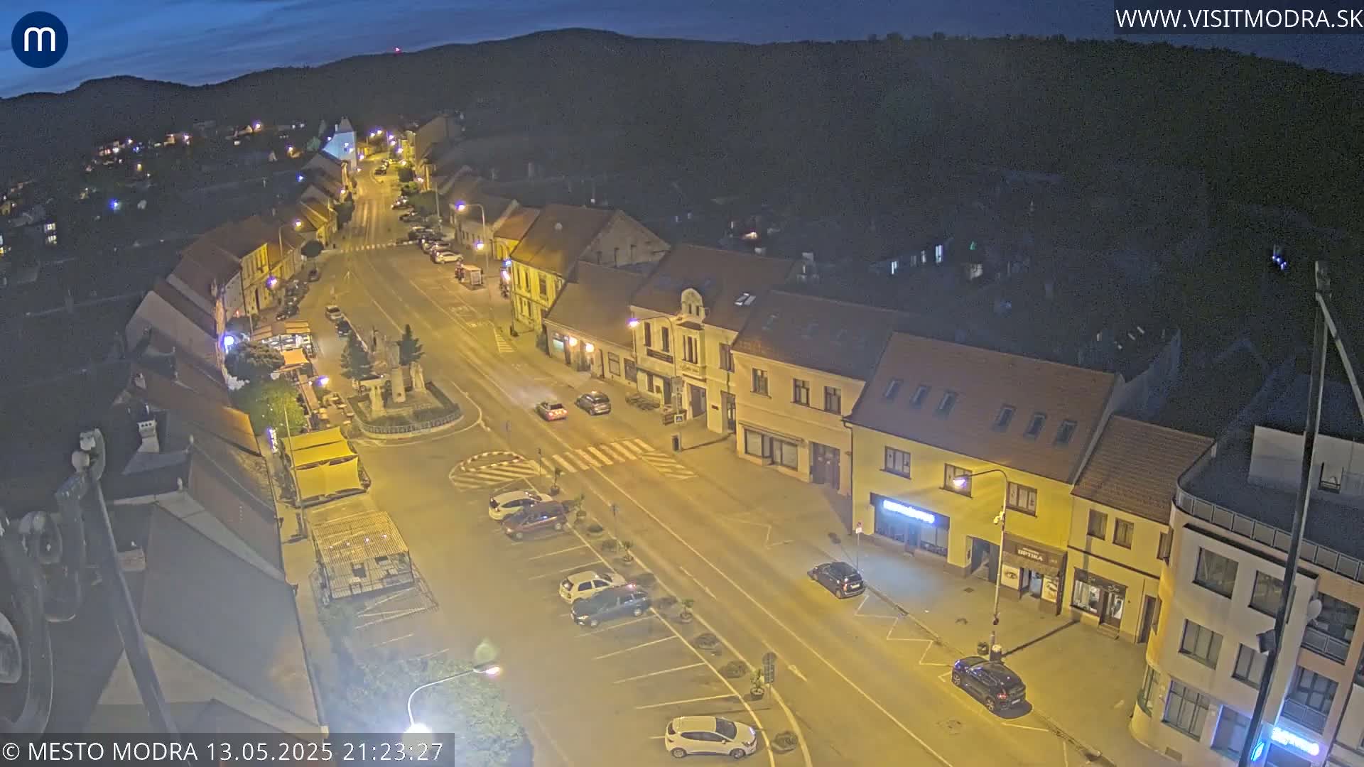 A high-angle, nighttime view of a town square with several cars parked in a lot and others driving on the road, under a dark sky.