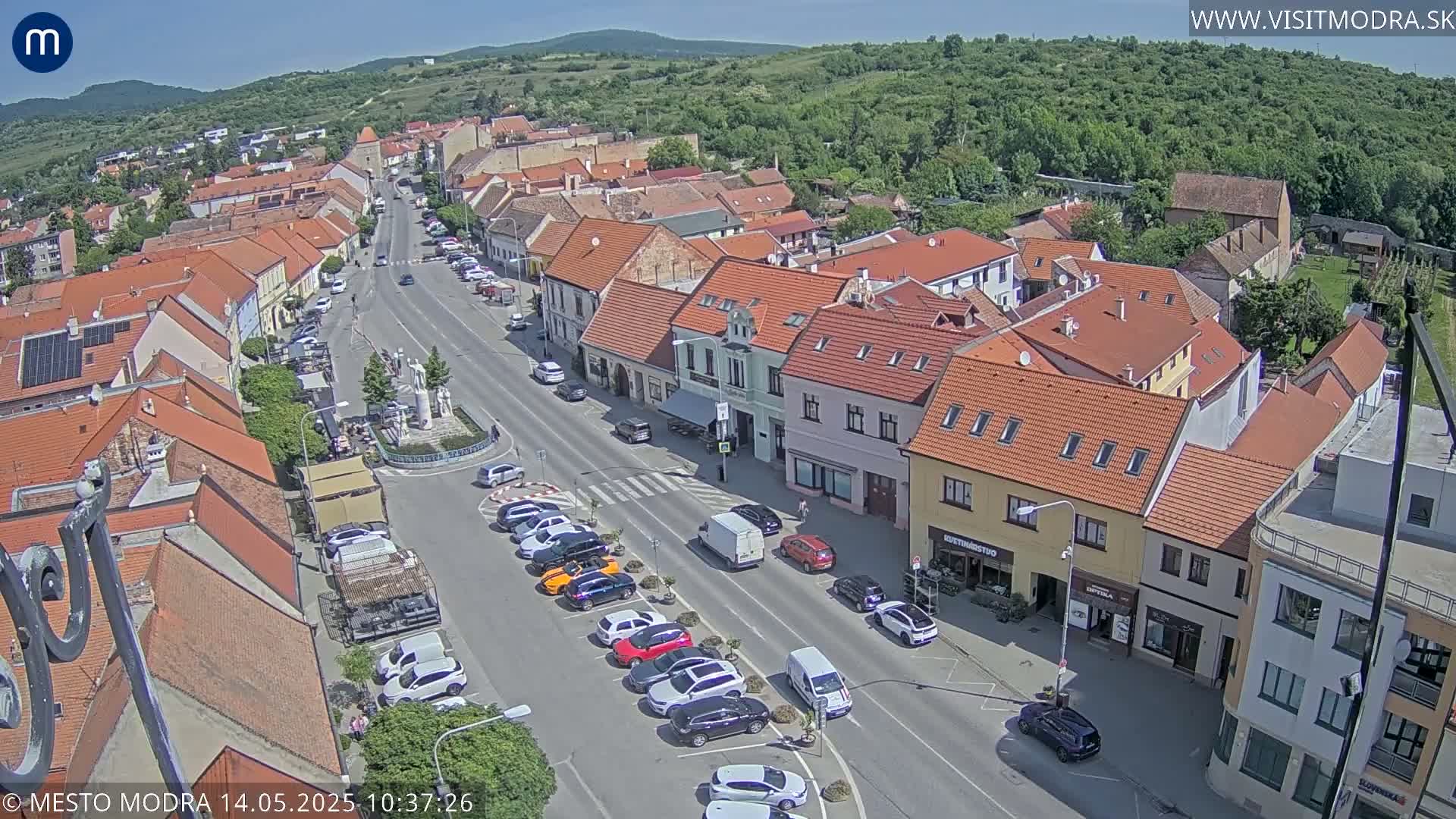 An aerial view shows a town square and street lined with buildings under a sunny sky.
